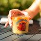 A hand holding a clear glass jar candle filled with orange wax, featuring a red coffee mug graphic and the text 'TECHYD DA!' on a dark wooden table with a blurred green garden background.